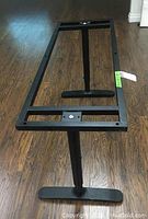 Full view of black rectangular metal table frame with central support and two feet on dark wooden floor.