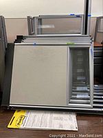 Stack of office cubicle partitions showing beige fabric panels with aluminum frames and glass window sections on one side.
