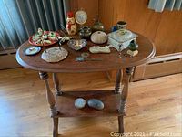 Full view of oval wooden table with turned legs and lower shelf displaying the animal figurines and assorted decorative items on its tabletop and decorative rocks below.
