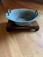 Side angle of verdigris patina metal vessel with brass handles positioned on wooden platform.