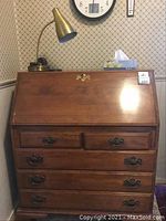 Front view of the wooden secretary desk showing the medium finish, drop-down front, and 5 drawers with brass handles.