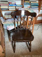 Dark wood chair with curved spindle back and contoured seat in front of two upholstered stools with pastel patchwork quilt seats, and a shelf with books behind.