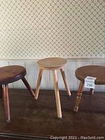 Three wooden milking stools arranged side by side on a wooden floor against a wallpapered wall, showing overall size and style.