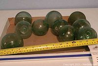 Set of ten green glass fishing floats arranged in a cardboard box with a measuring tape showing size approximately 4 inches each.