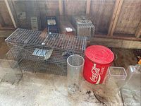 Image of multiple wire mesh animal traps, a large red Coca Cola Classic metal can, and three clear glass vases arranged on a concrete floor.