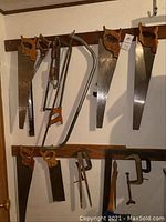 Multiple saws with wooden handles hanging on a wall rack with clamps and other tools below