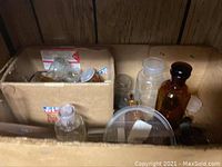 Photo showing assorted glass bottles and jars in cardboard box, some amber glass and some clear