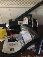Metro Omnivac black vacuum cleaner main unit with power cord, shoulder strap, and instruction manual on table.