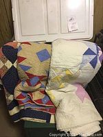 Photo showing three vintage quilts folded together on top of a green surface with white cabinet behind them.