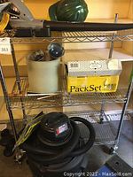 Photo showing chrome rack shelving with the blower on the top shelf alongside hardhats and packing box. Shop vac with hose sits on bottom shelf.
