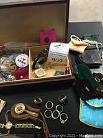 Overall view of wooden box with selection of watches, rings, pins, pouches and necklace on black surface