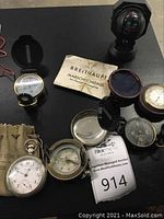 Top view showing multiple vintage compasses including Breithaupt Marsch-Kompass and a vintage pocket watch with cloth pouch on a black surface.