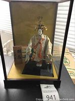 Full view of Japanese doll inside glass display case showing doll, wooden plaque, and postcard inside case