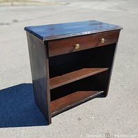 Front angle of wooden nightstand showing drawer with brass knobs and two open shelves below.