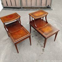 Pair of wooden end tables with brown leather tops and gold embossing, placed outdoors on pavement.