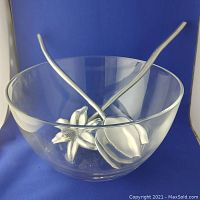 Clear glass salad bowl with pair of long-handled metal serving tongs inside, set against blue background showing profile view of tongs and flower-shaped heads.