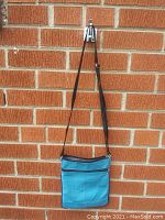 Front view of turquoise leather tote bag hanging against brick wall showing stitching details and zippered closure