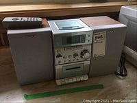 Front view of Sony compact stereo system showing two speakers, stereo unit with tape deck, CD player, and tuner controls, and remote control on top of speaker.