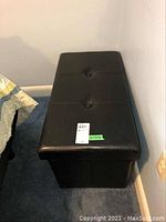 Photo of full black rectangular storage bench with tufted button detailing on top and hinged lid closed, positioned near corner of a room with blue carpet.