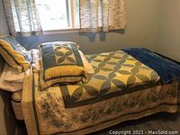 A bed made up with a quilt that has a large geometric pattern in yellow, blue, and cream, with a floral border. Two matching pillow shams are on one side of the bed and a solid dark blue throw blanket is folded at the foot.