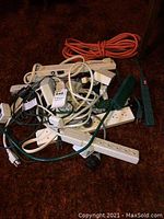 Photo showing a pile of various extension cords, power bars, and a Noma timer on a dark carpeted floor.