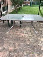 Full image of a rectangular patio table with a glass top and white metal base placed on a paved patio area outdoors.
