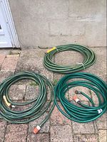 Three garden hoses placed outside on pavement near wall, two are green YarkWorks hoses with quick release connectors, the third hose is a different green hose without quick release connectors.