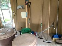 Photo of yard tools including rakes, shovels, broom, squeegee and garbage cans placed indoors