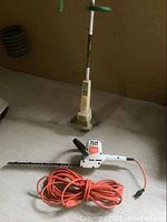 Photo showing electric weed trimmer with tall vertical handle and base, and in front the hedge trimmer and coiled orange extension cord on concrete floor.