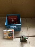 Blue plastic milk crate with red metal Ontario 1977 first aid kit and car jack handle in front, box of booster cables partially visible