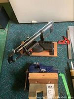 Full view of Jobmate mitre saw mounted on wood platform with protractor scale and safety guard in elevated position beside the blue Mastercraft vice mounted on a wooden base sitting on green carpet, along with measuring tools.