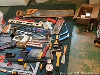 Wide view of assorted hand tools laid out on floor including saws, levels, scrapers, hand drill and clamps.