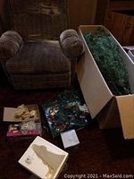 Photo of the artificial Christmas tree branches in the box positioned upright next to a brown armchair, along with assorted Christmas lights and some ornaments scattered nearby on the floor.