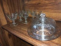 Six etched glass dessert or wine glasses arranged on wood shelf next to glass butter dish with domed lid