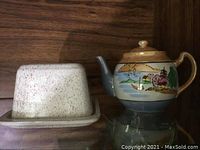 Ceramic lidded butter dish with speckled glaze and a small painted teapot on a wooden shelf.