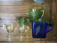 Assorted pressed glassware displayed on wooden shelf, includes green and blue pitchers and cream and sugar sets in green and clear glass.