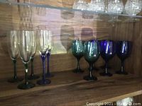 Set of 5 clear champagne glasses and 6 blue and green wine glasses in a wood shelf.