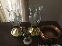 Pair of etched glass candleholders with brass bases, small Goebel figurine, and small copper cauldron grouped on dark surface.