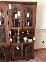 Front view of cabinet showing upper and lower glass paneled doors, decorative wood trim, interior contents, and brass handles.