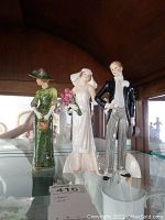 Three figurines displayed on glass shelf: green-dress Edwardian lady, bride, groom