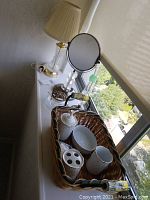 Full view of the wicker basket with ceramic bathroom items and round vanity mirror on stand, glass table lamp on the side