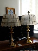 Pair of matching brass table lamps with scalloped, translucent, shell-like lampshades on wooden surface in front of a window.