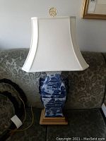 Full view of the blue porcelain table lamp with white fabric shade on a patterned sofa showing the whole lamp and base.