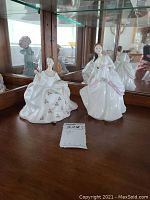 Two Royal Doulton figurines placed on a wooden surface in front of a mirror; one is Carol HN 2961 holding a red flower, dressed in white with gold floral details; the other is My Love HN 2339 with a white and pink dress holding her skirt