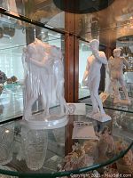 The first photo shows two white porcelain Greek-style figurines inside a glass cabinet. On the left is a multi-figure group of three women, and on the right is a nude male figure on a rectangular base.