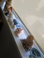 Long view of the shelf displaying various large seashells, blue and white ceramic box, decorative candles, and the shell-embedded lamp with carved fairy inside on the right side.