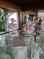 Glass shelf with multiple porcelain figurines, focusing on two pink dress Dresden pieces in front