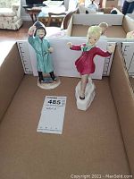 Two Royal Worcester porcelain figurines, one in blue coat and one in pink coat, standing in a cardboard box.