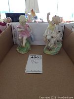 Photo showing both Royal Worcester figurines side by side in a cardboard box. One girl in pink dress holding shoes and another girl in yellow dress playing with a dog.