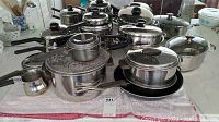 Photo showing various stainless steel pots and pans arranged on a towel, with black handles and lids. Includes a colander and small saucepans.
